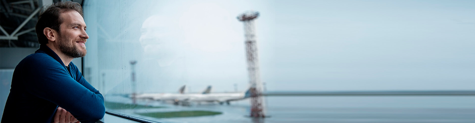Suscripcion fondos de inversion - Hombre con una camiseta azul sonriendo y mirando por la ventana en un aeropuerto Suscripcion fondos de inversion - Hombre con una camiseta azul sonriendo y mirando por la ventana en un aeropuerto