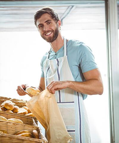 Seguro Multirriesgo de Comercio - Panadero joven sonriendo mientras se encuentra cogiendo pan en su negocio