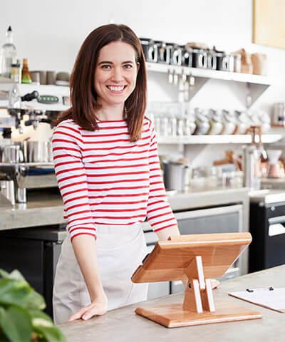 Ventajas TPV Fijo - Mujer joven con camiseta de rayas rojas y blancas, dueña de una cafeteria sonriendo detras de la barra
